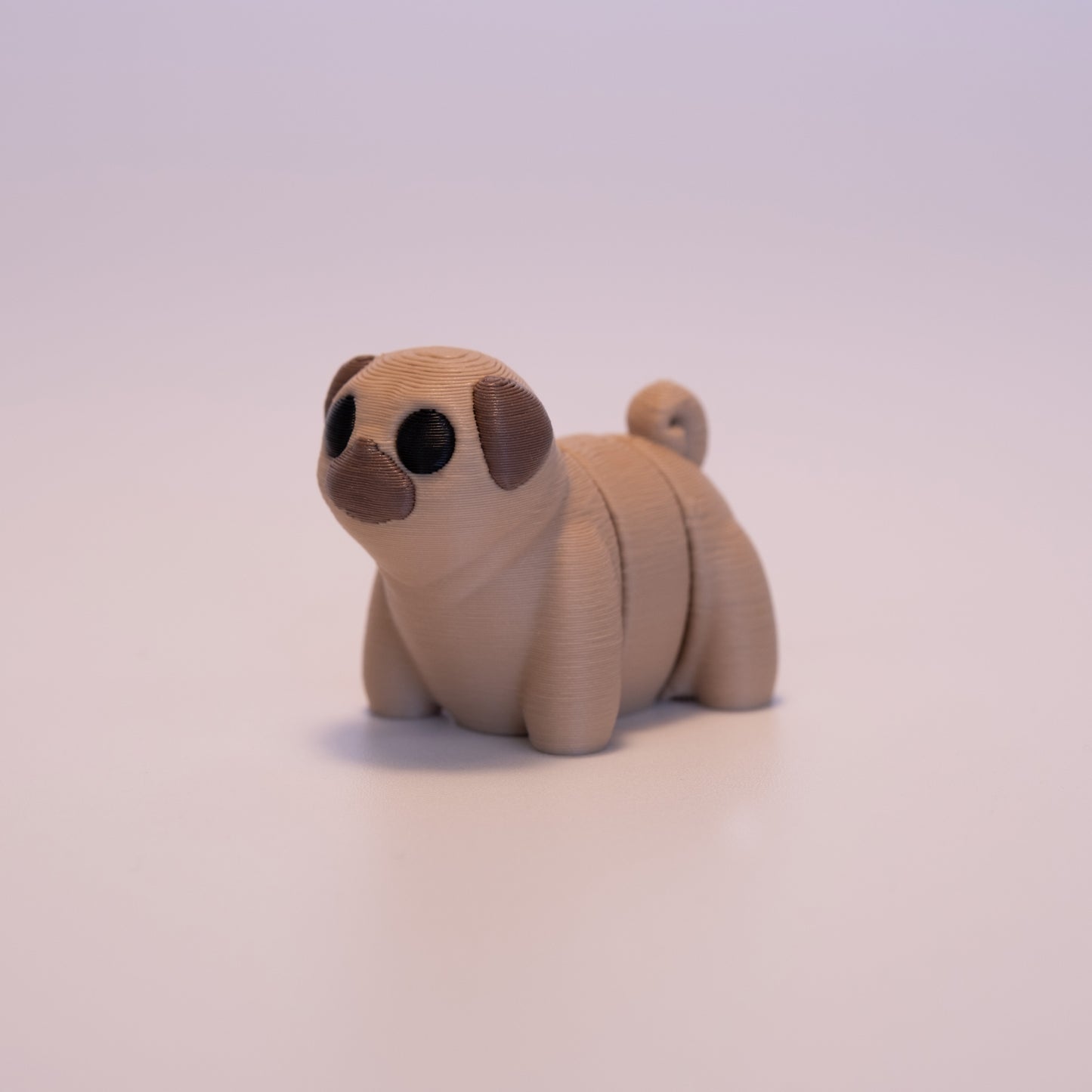 A 3D Printed Pug figurine standing on its 4 legs on an off white background. The Pug is mostly tan with brown ears, brown nose, and round black eyes.
