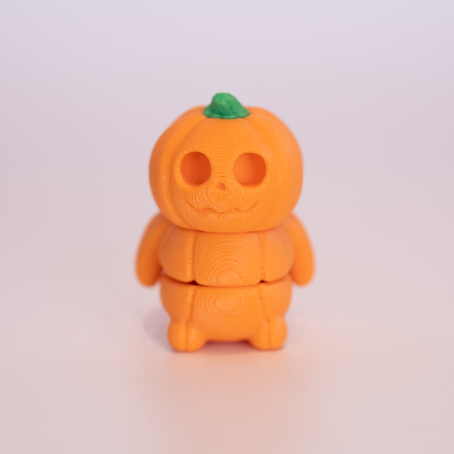 A 3D printed standing Pumpkin on an off white background. The Pumpkin is humanoid in shape. It is almost entirely orange with a green stem.