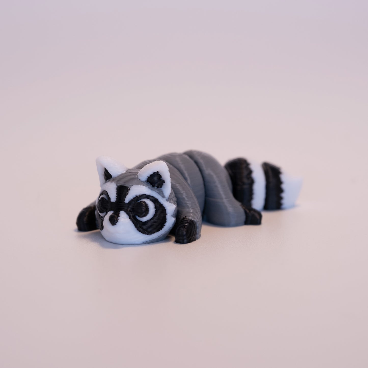 A 3D printed Raccoon figurine laying on its stomach over an off white background. The figurine is mostly grey with black paws, white scleras, and black iris/pupils looking up. The Raccoon also has white and black: ears, face, and striped tail.