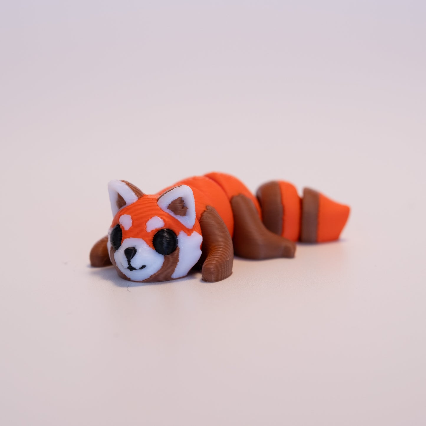 A 3D printed Red Panda figurine laying on its stomach over an off white background. The figurine is mostly dark orange with warm brown limbs, round black eyes, and a black nose and mouth. The Red Panda also has a dark orange and warm brown striped tail, white and brown ears and white and brown accents on the face.