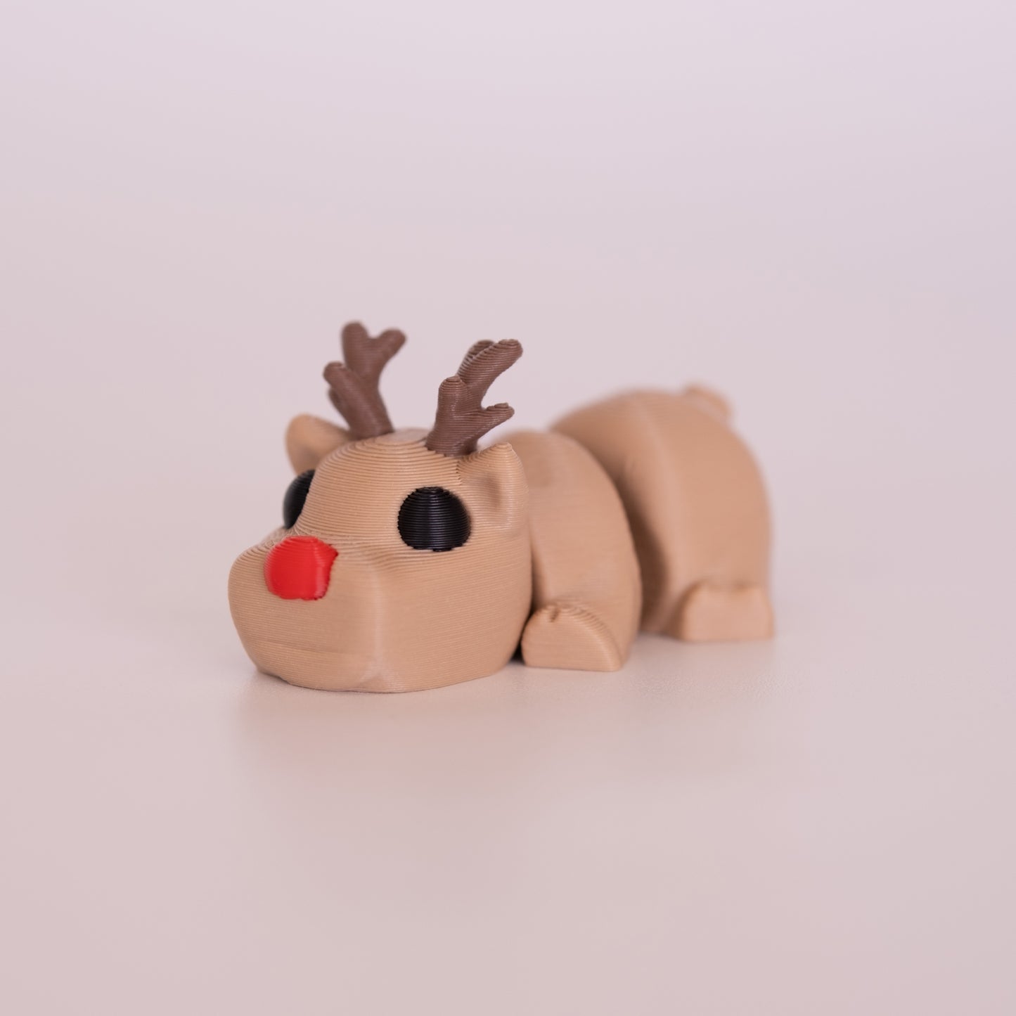 A 3D printed Rudolph figurine laying on its stomach over an off white background. The figurine is mostly tan with round black eyes, brown antlers, and a red nose.