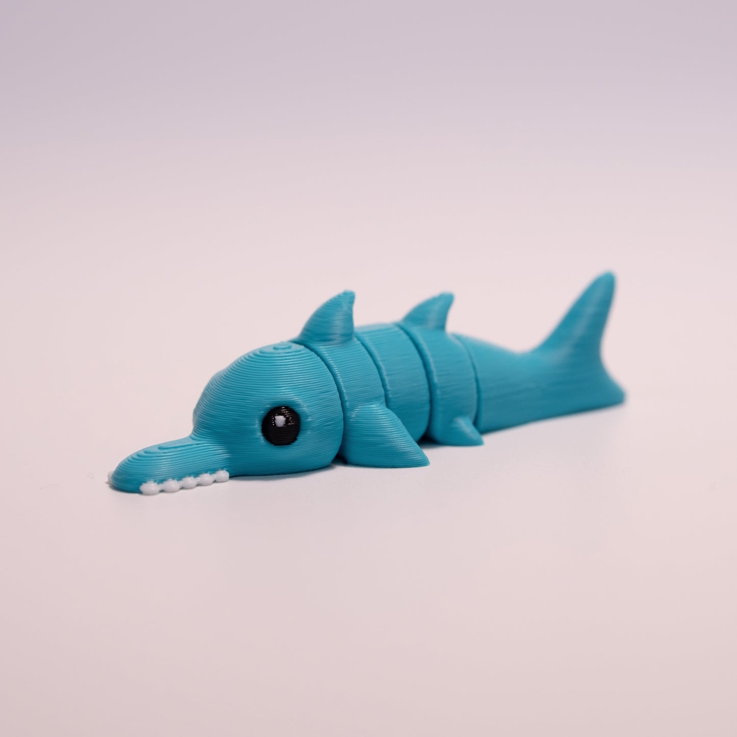 A 3D printed Sawshark figurine laying on its stomach over an off white background. The figurine is mostly teal, round black eyes with a small white accent dot in each eye, and white teeth.