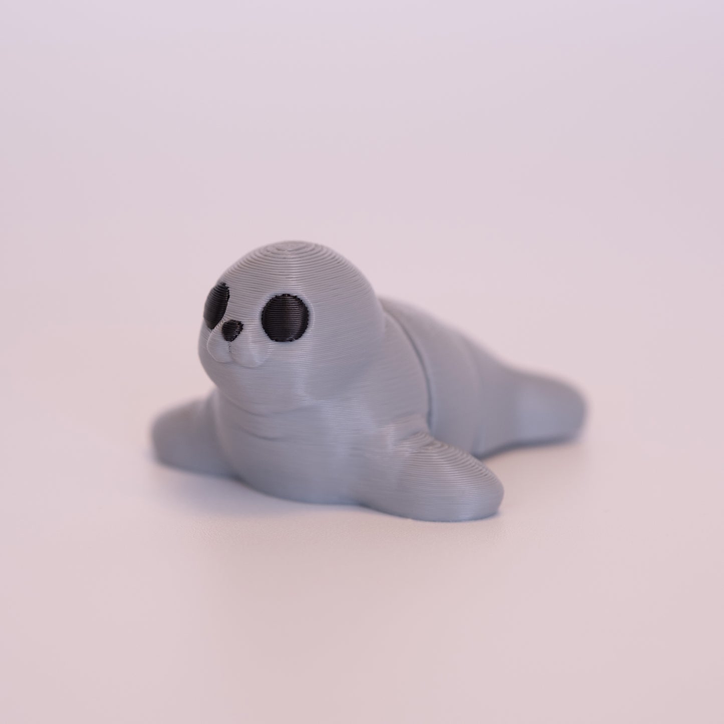 A 3D printed Seal figurine laying on its stomach over an off white background. The figurine is entirely light grey with round black eyes and a black nose.