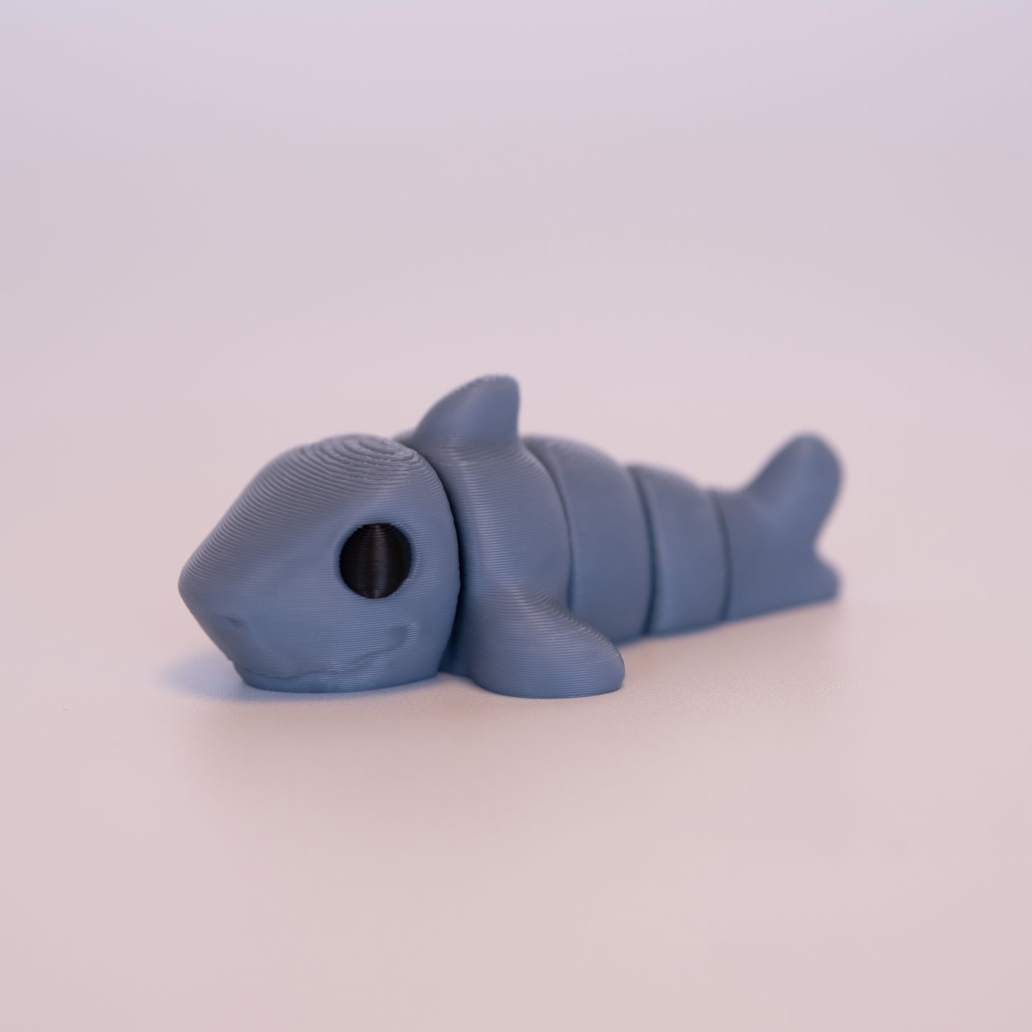 A 3D printed Shark figurine laying on its stomach over an off white background. The figurine is entirely blue-grey with round black eyes.