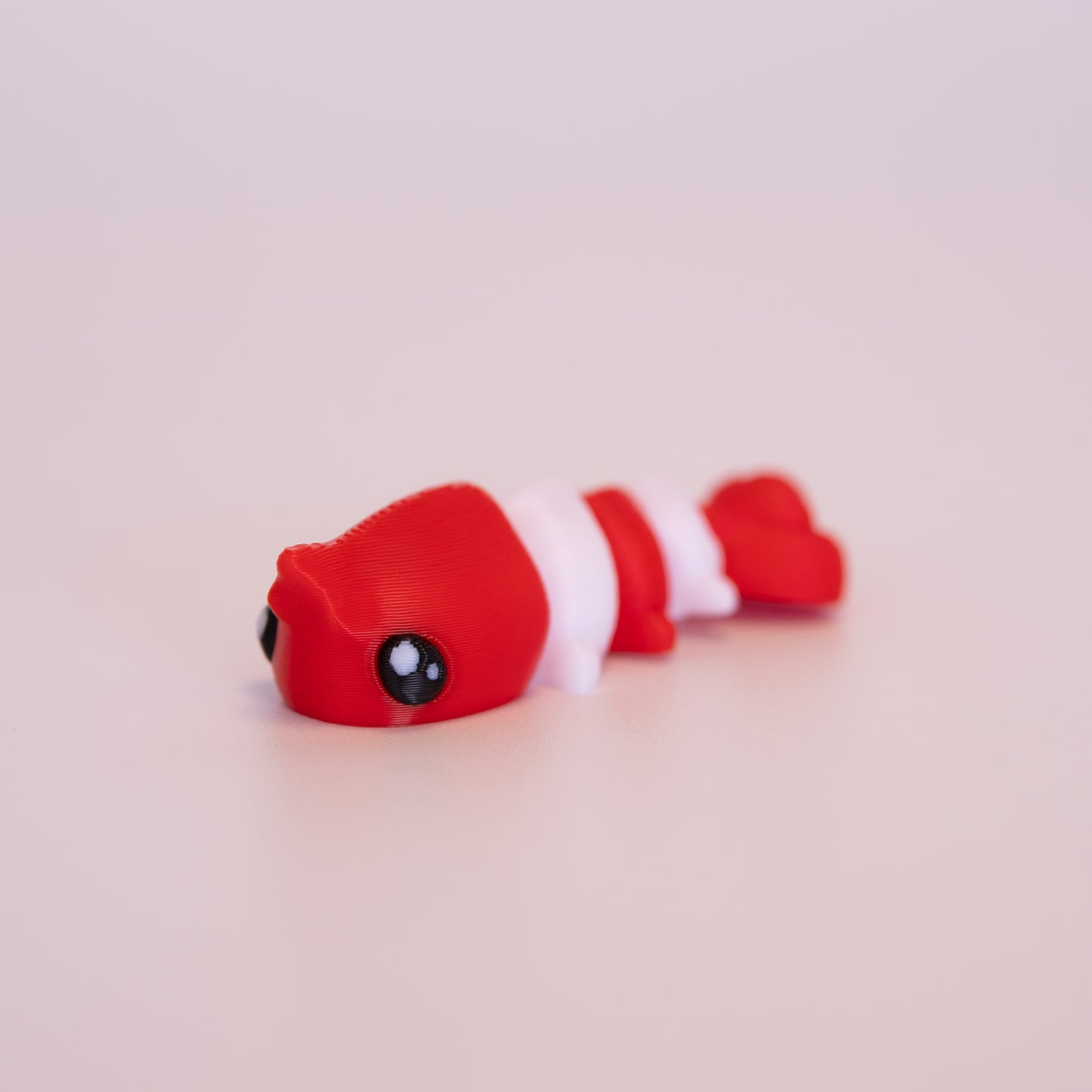 A 3D printed Shrimp figurine laying flat on an off white background. The figurine is red and white striped and has round black eyes with two white accent dots in each eye.
