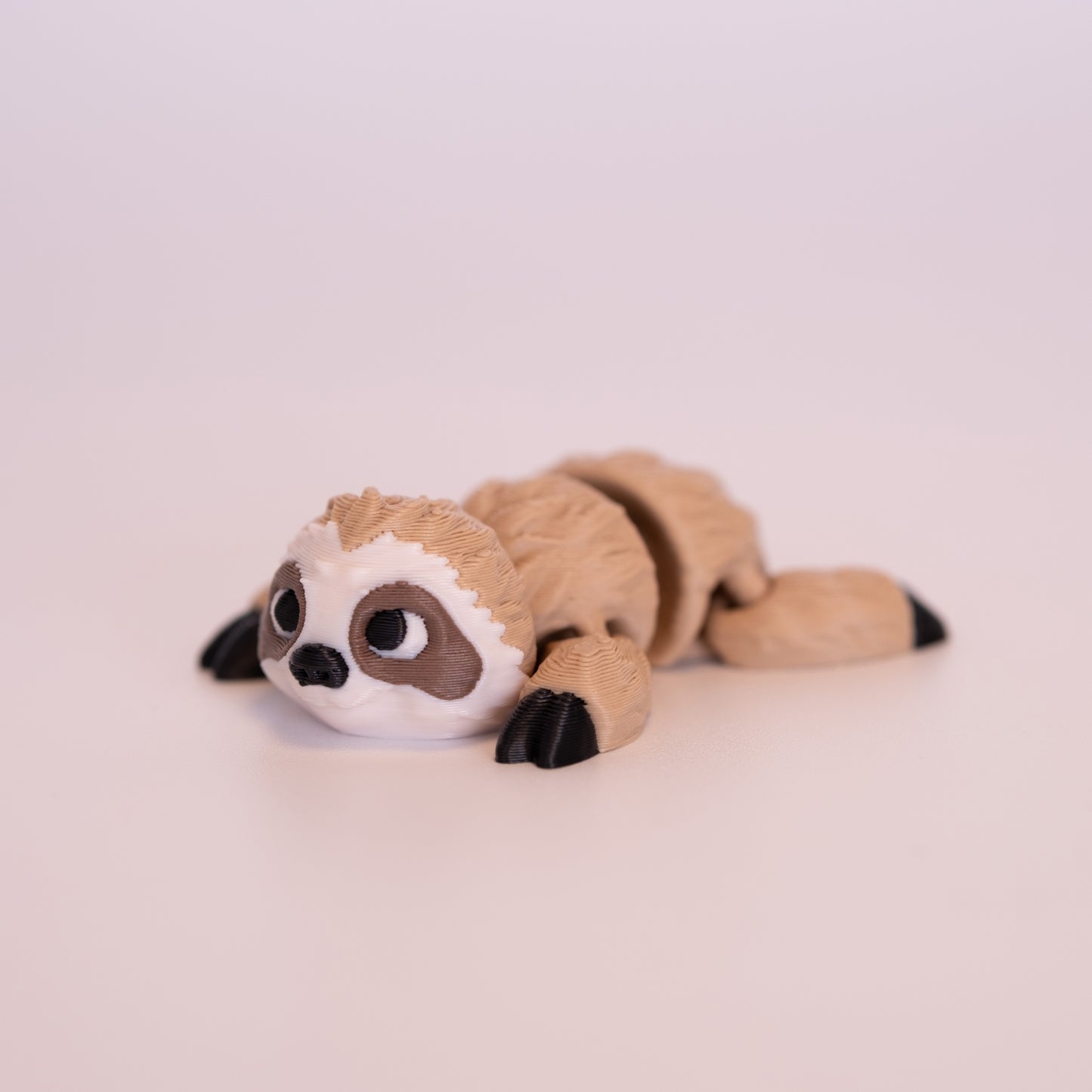 A 3D printed Sloth figurine laying on its stomach over an off white background. The figurine is mostly tan, black nails, cream face, brown accent around the eyes, white scleras, and big black pupils looking forward.