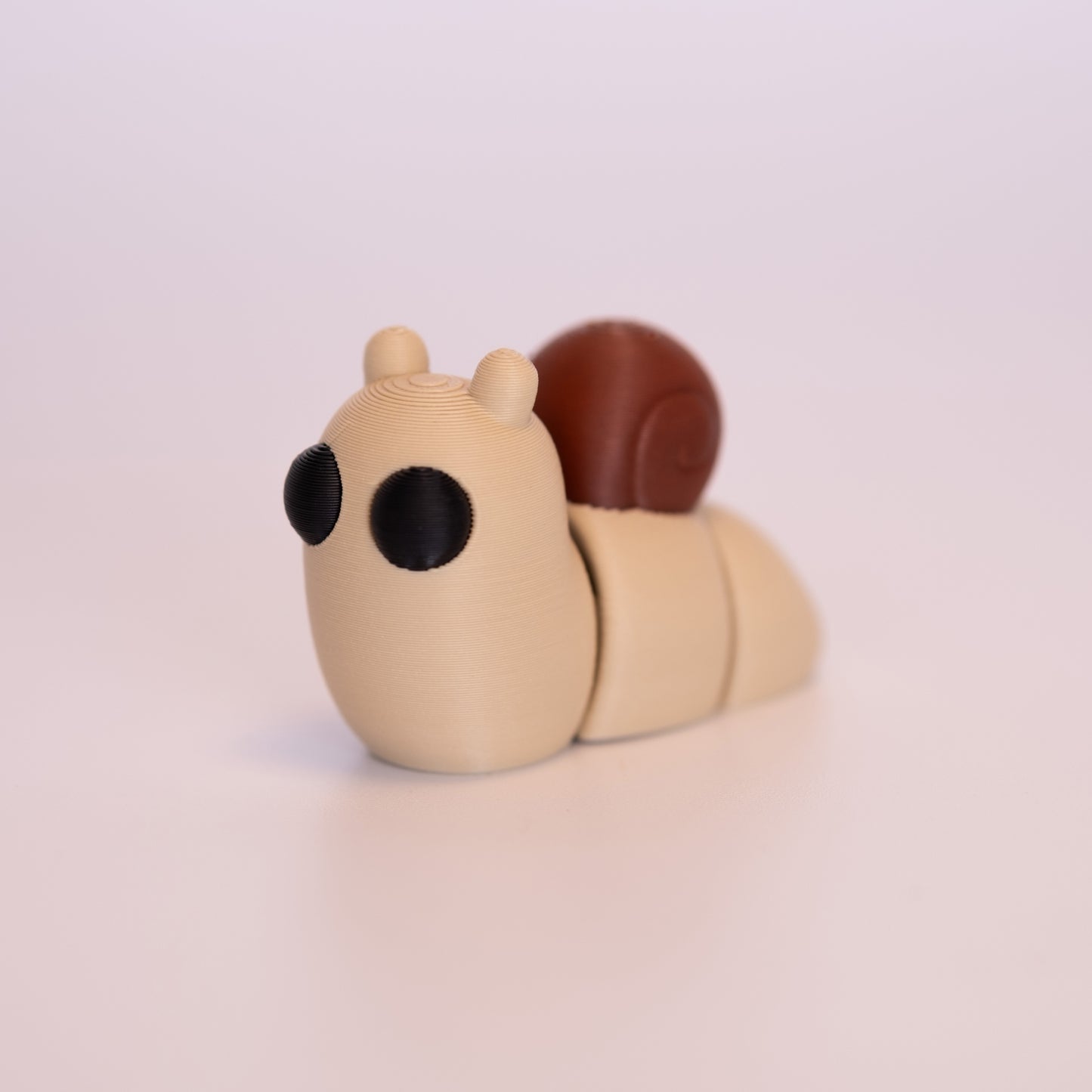 A 3D printed Snail figurine laying on an off white background. The figurine is mostly tan, round black eyes, and a brown shell.