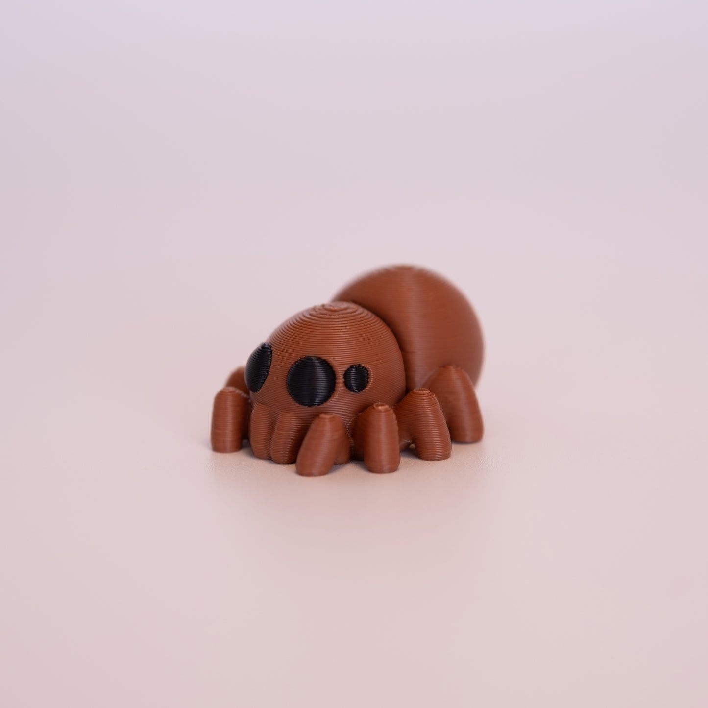A 3D Printed Spider figurine standing on its 8 legs on an off white background. The Spider is mostly a warm brown with round black eyes.
