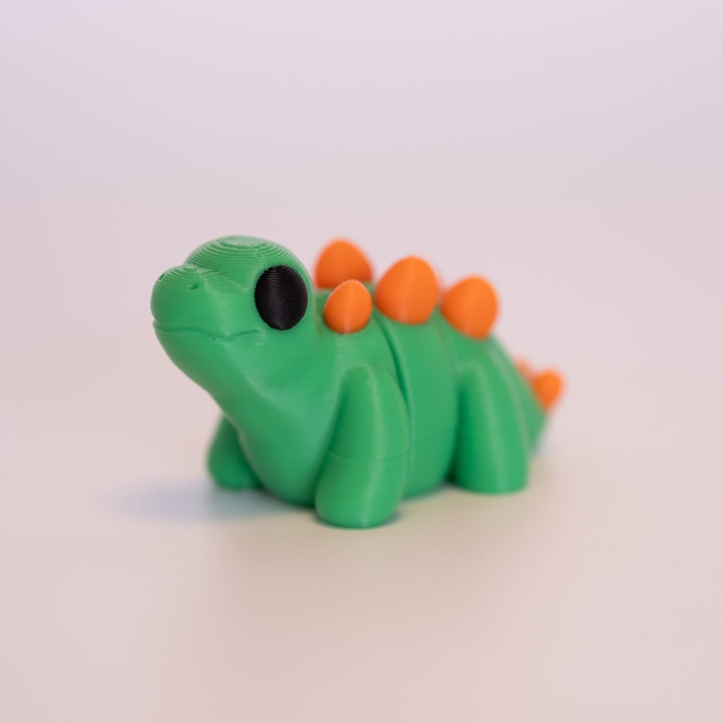 A 3D Printed Stegosaurus figurine standing on its 4 legs on an off white background. The Stegosaurus is mostly green with round black eyes, and orange accents for the “spikes”.