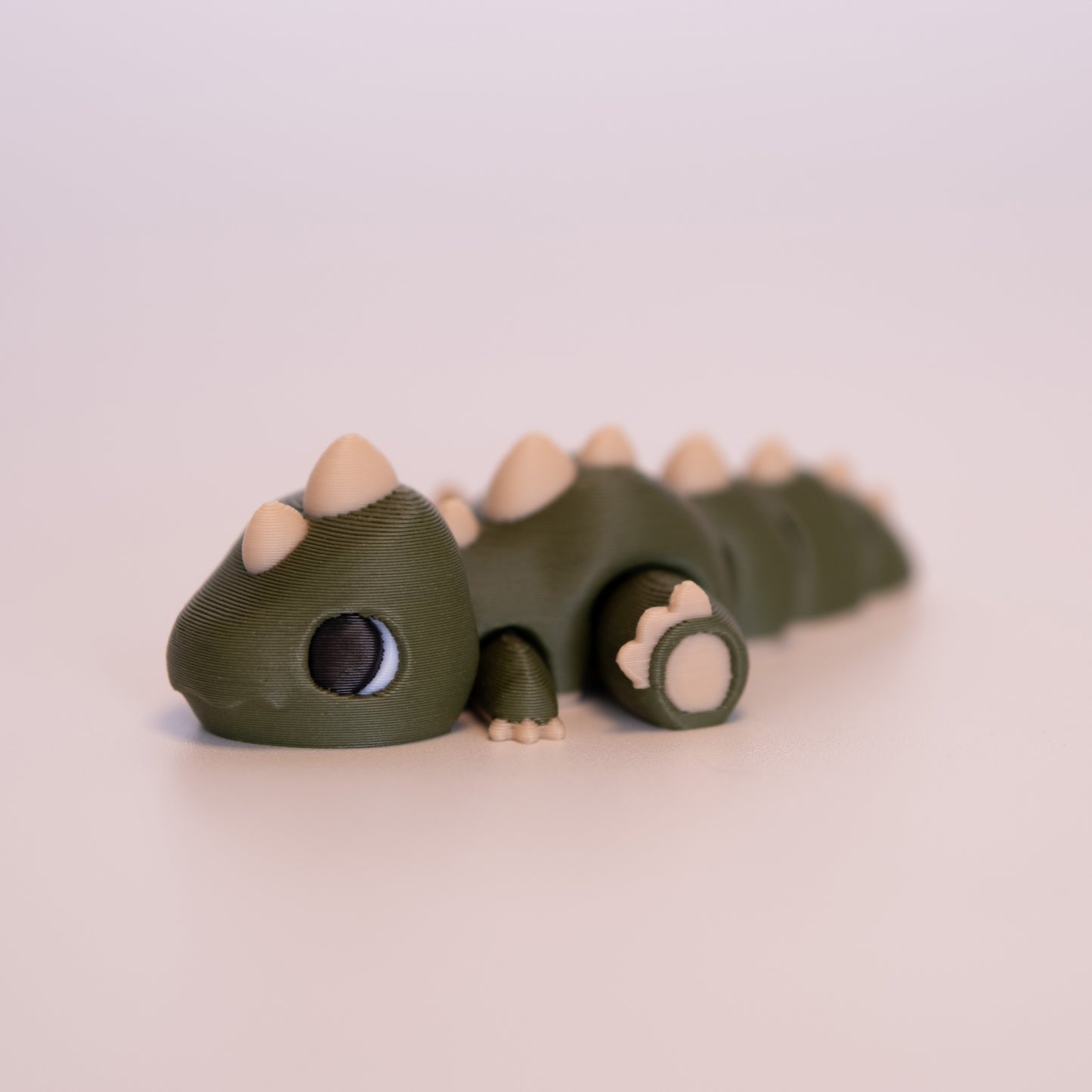 A 3D printed T-Rex figurine laying on its stomach over an off white background. The figurine is mostly dark green with cream accents, white scaleras, and large black pupils looking forward.