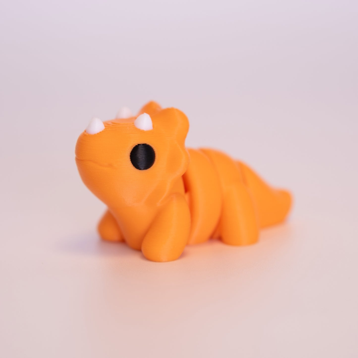 A 3D Printed Triceratops figurine standing on its 4 legs on an off white background. The Triceratops is mostly orange with round black eyes, and white horns.