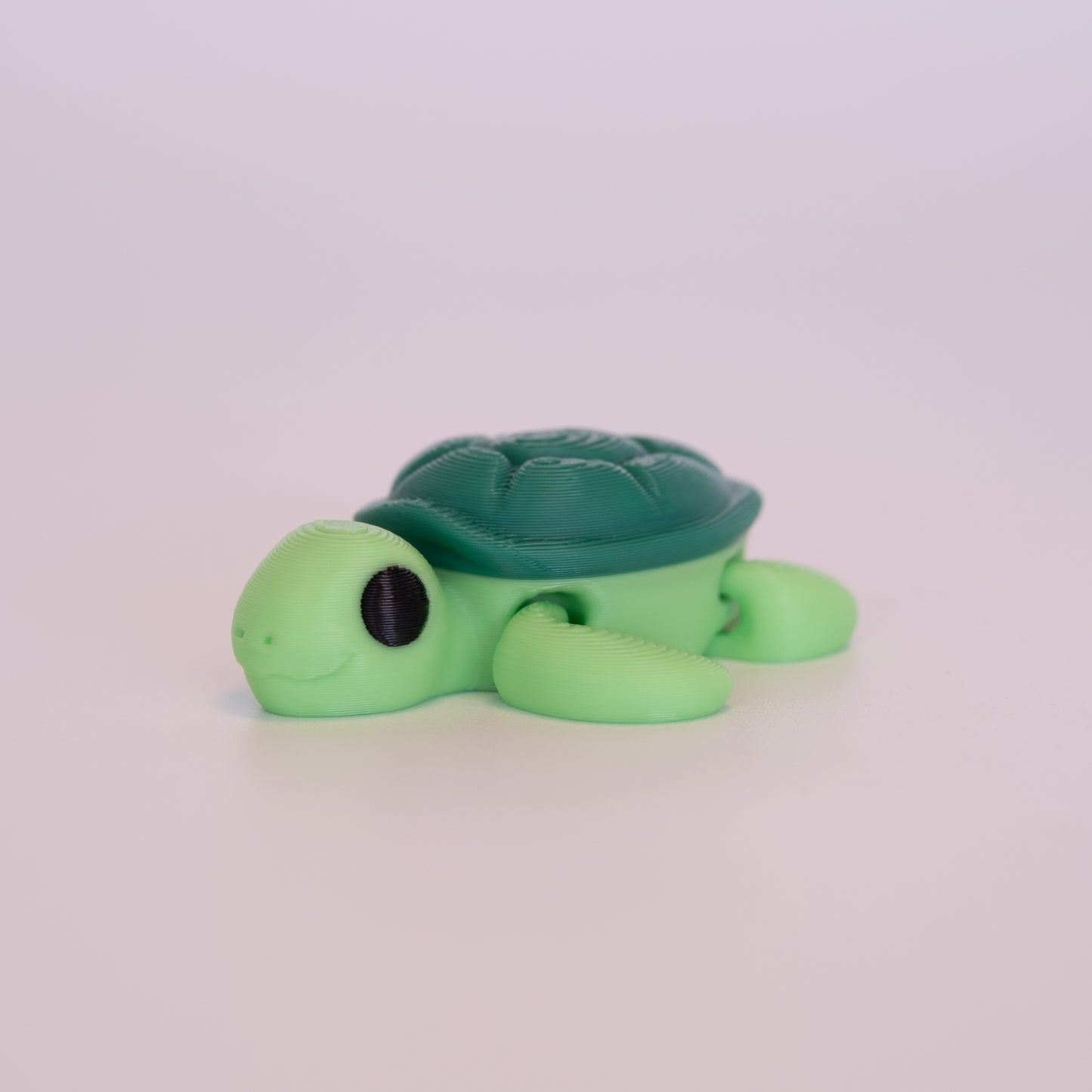 A 3D printed Sea Turtle figurine laying on its stomach over an off white background. The figurine has a light green body, dark green shell, and black eyes.