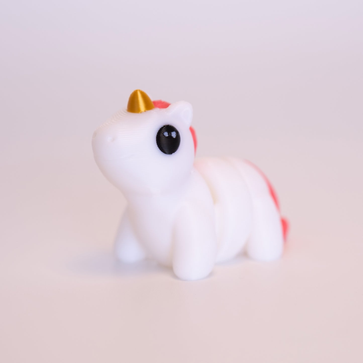 A 3D Printed Unicorn figurine standing on its 4 legs on an off white background. The Unicorn is mostly white, with a gold horn, pink mane and tail, and round black eyes with 2 small white accent dots in each eye.