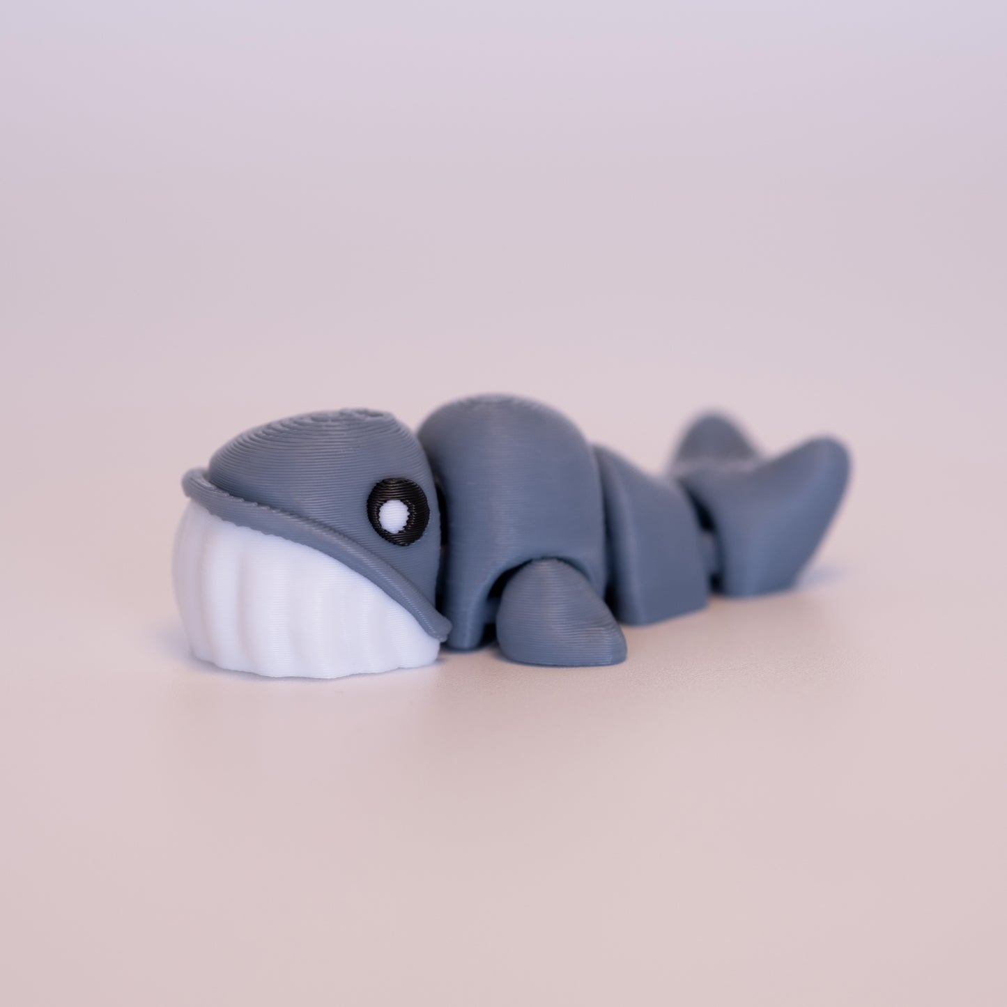 A 3D printed Blue Whale figurine laying on its stomach over an off white background. The figurine is mostly blue-grey, round black eyes with a white dot accent in each eye, and white bottom half of the face.