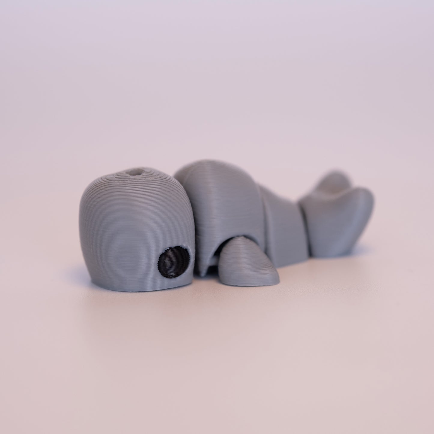 A 3D printed Sperm Whale figurine laying on its stomach over an off white background. The figurine is entirely light grey with round black eyes.