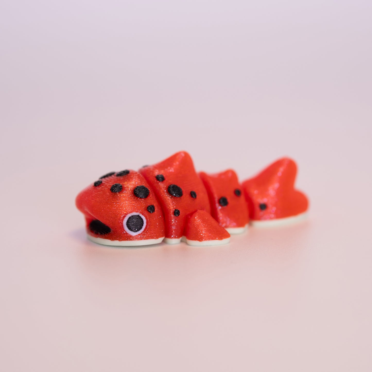 A 3D printed Watermelon Whale Shark figurine laying on its stomach over an off white background. The figurine is mostly shimmery red, black spotted accents, black mouth, cream bottom, white scaleras, and big black pupils.
