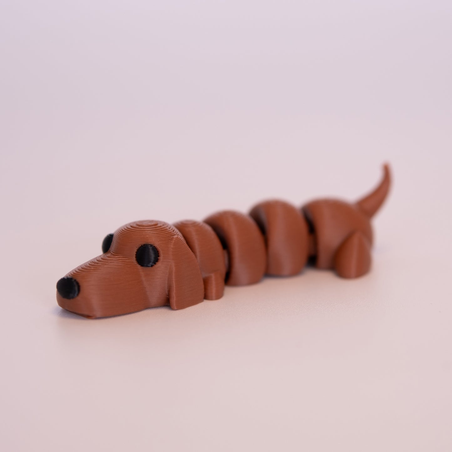 A 3D printed Wiener Dog figurine laying on its stomach over an off white background. The figurine is entirely a warm brown with round black eyes and a black nose.