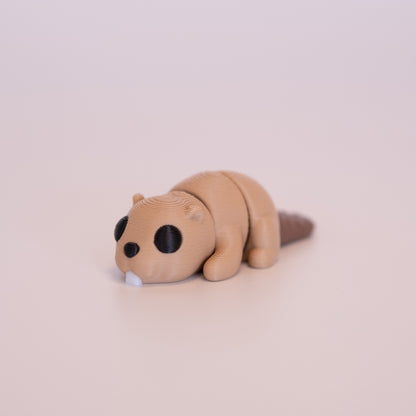 A 3D printed Beaver figurine laying on its stomach over an off white background. The figurine is mostly tan with round black eyes, white buck teeth, and a brown tail.