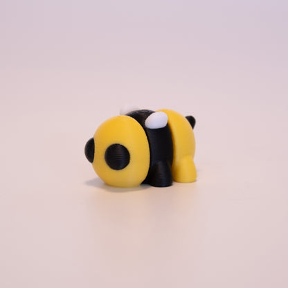 A 3D Printed Bee figurine standing on its 4 legs on an off white background. The Bee’s body has alternating colors of yellow and black, with round black eyes, white wings, and a black stinger. 