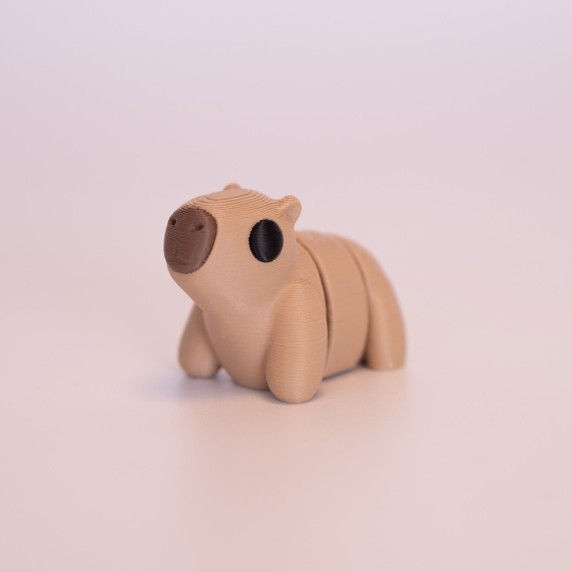 A 3D Printed Capybara figurine standing on its 4 legs on an off white background. The Capybara is mostly tan with round black eyes, and a brown nose. 