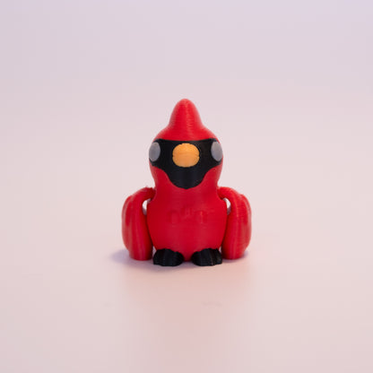A 3D printed Cardinal sitting upright on an off white background. The Cardinal is mostly red with grey eyes, orange beak, black feet, and black accents on its face.