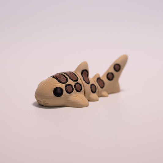 A 3D printed Catshark figurine laying on its stomach over an off white background. The figurine is mostly tan with round black eyes and brown and black patterned accents.