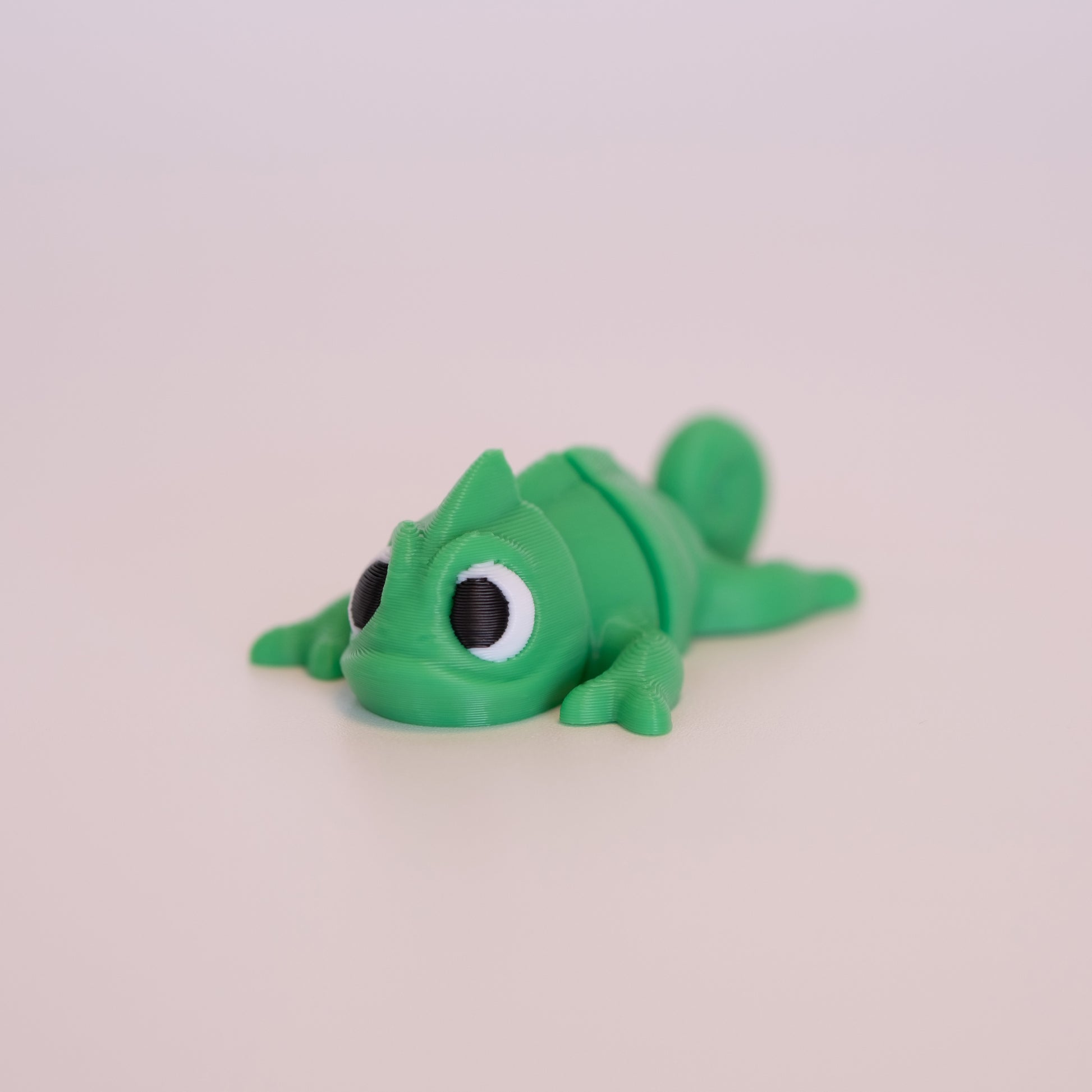 A 3D printed Chameleon figurine laying on its stomach over an off white background. The figurine is mostly green with round black eyes,white scleras, and black iris/pupils looking forward.