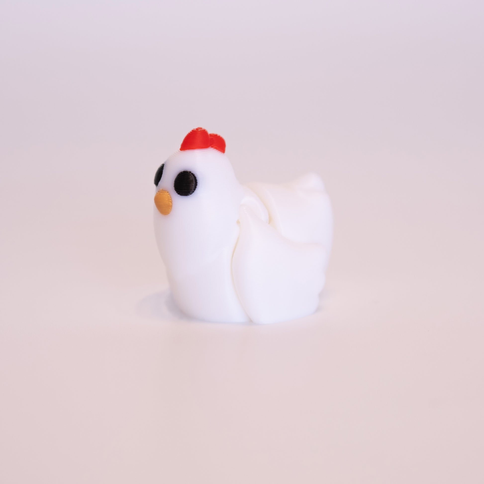 A 3D printed Chicken sitting upright on an off white background. The Chicken is mostly white with round black eyes, gold beak, and red accent on top of its head.