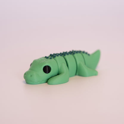 A 3D printed Crocodile figurine laying on its stomach over an off white background. The figurine is entirely green with round black eyes, and dark green accents.