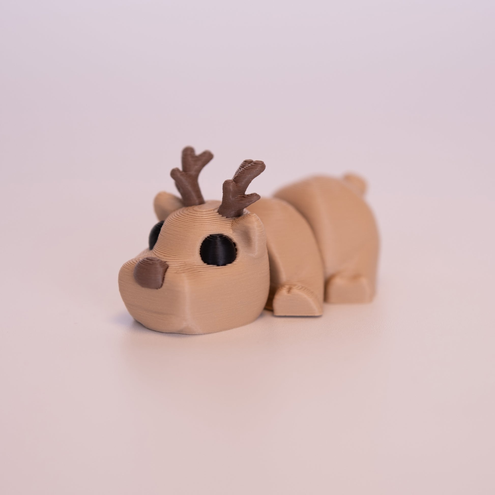 A 3D printed Deer figurine laying on its stomach over an off white background. The figurine is entirely brown with round black eyes, brown nose, and brown antlers.