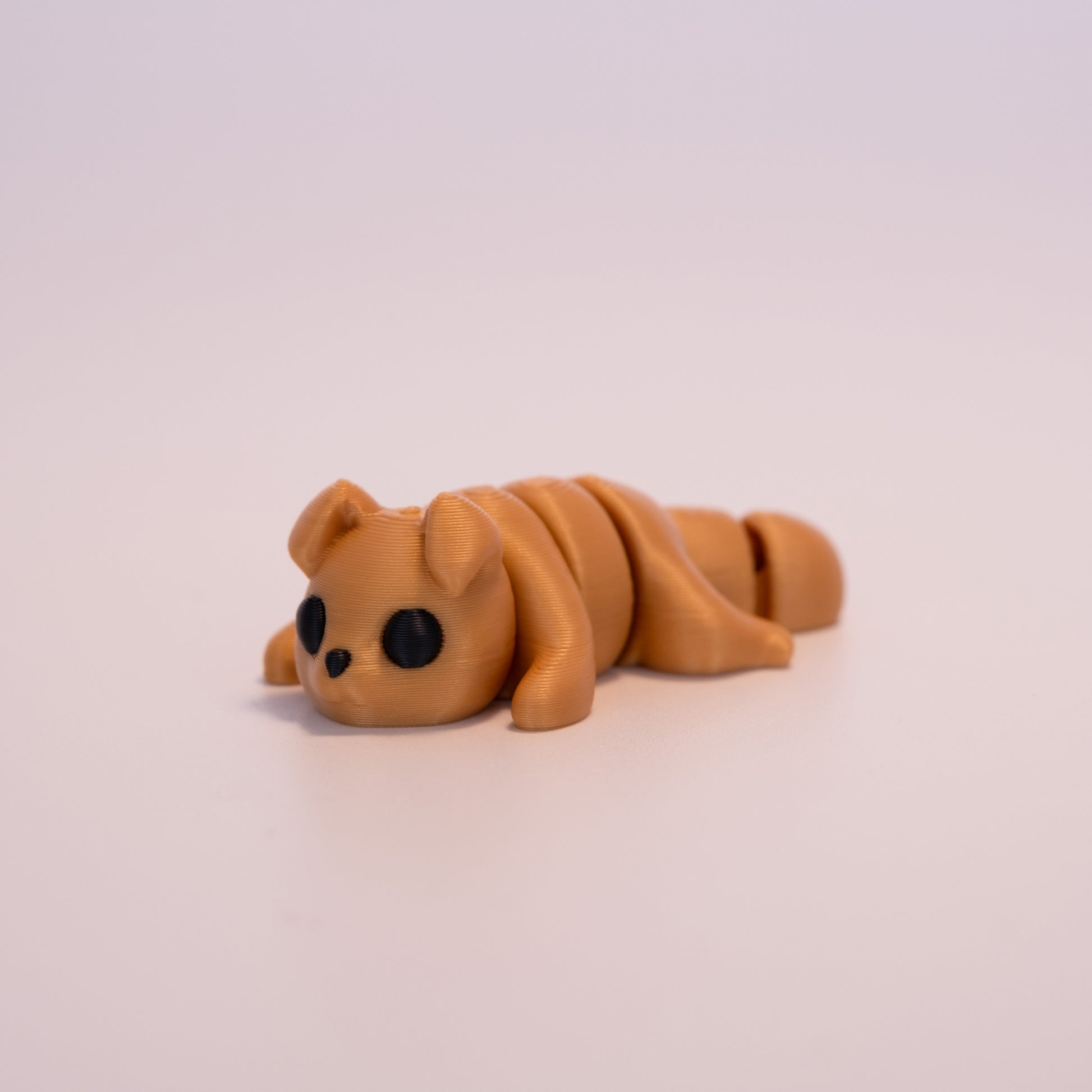 A 3D printed Gold Dog figurine laying on its stomach over an off white background. The figurine is entirely gold with round black eyes and a black button nose.