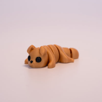 A 3D printed Gold Dog figurine laying on its stomach over an off white background. The figurine is entirely gold with round black eyes and a black button nose.