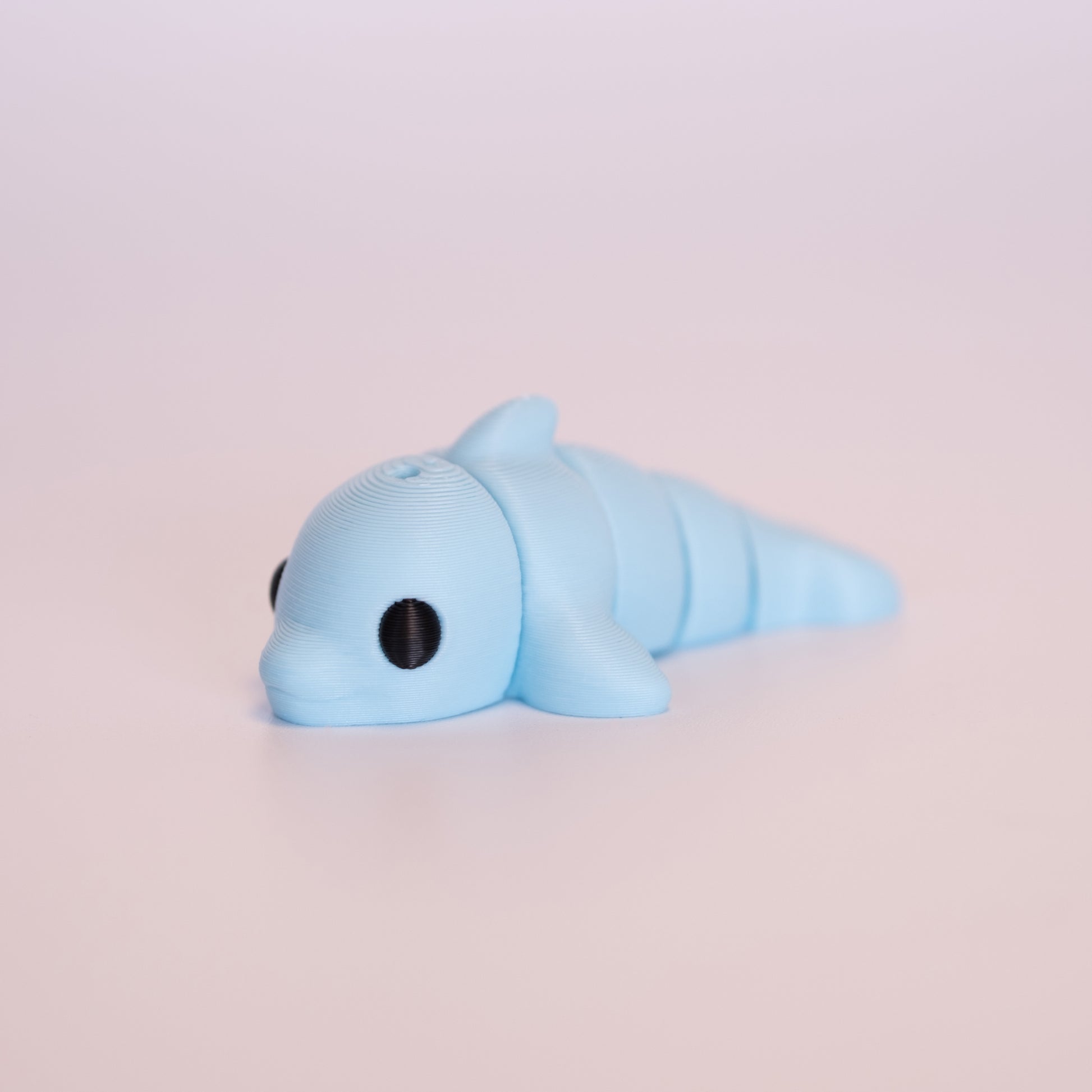A 3D printed Dolphin figurine laying on its stomach over an off white background. The figurine is entirely light blue with round black eyes.