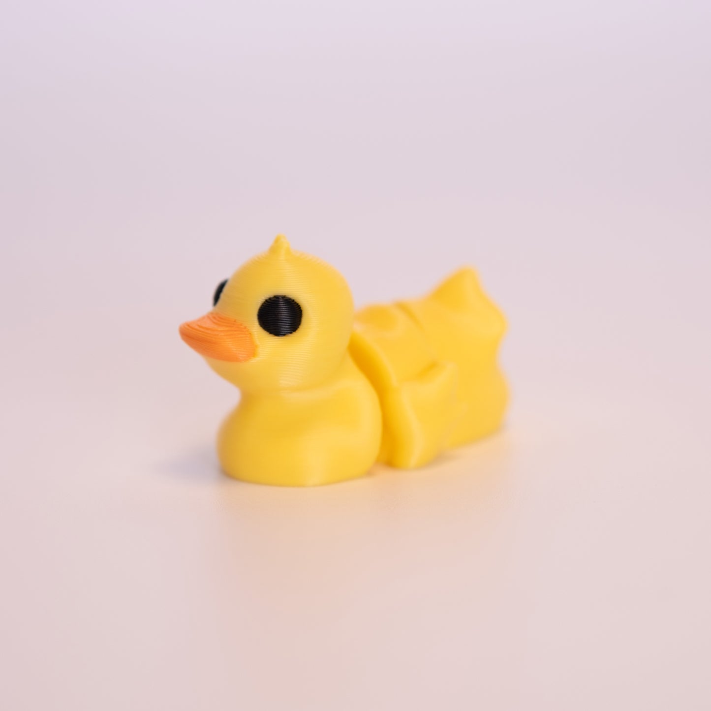 A 3D printed Duck sitting on an off white background. The Duck is mostly yellow with round black eyes, and an orange beak. 