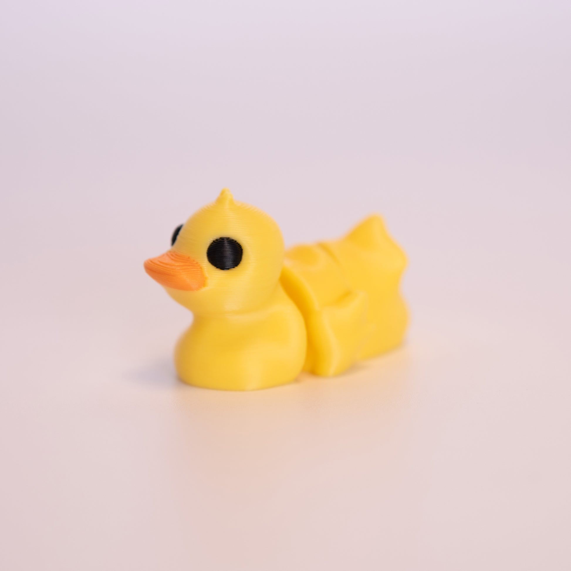 A 3D printed Duck sitting on an off white background. The Duck is mostly yellow with round black eyes, and an orange beak. 