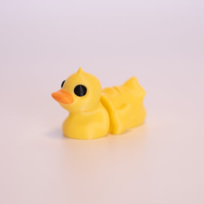 A 3D printed Duck sitting on an off white background. The Duck is mostly yellow with round black eyes, and an orange beak. 
