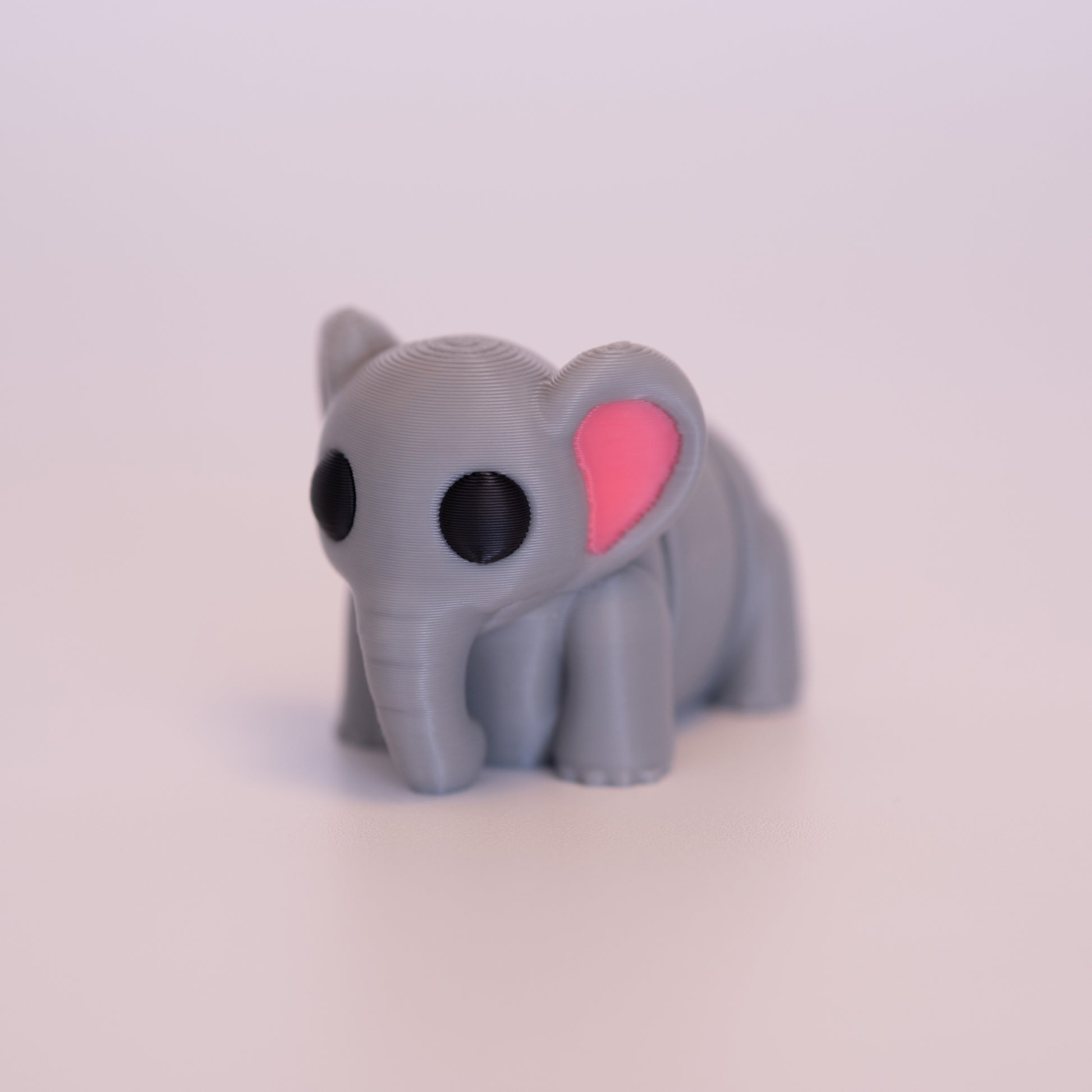 A 3D Printed Elephant figurine standing on its 4 legs on an off white background. The Elephant is mostly grey with round black eyes, and pink accents in the inner ears. 