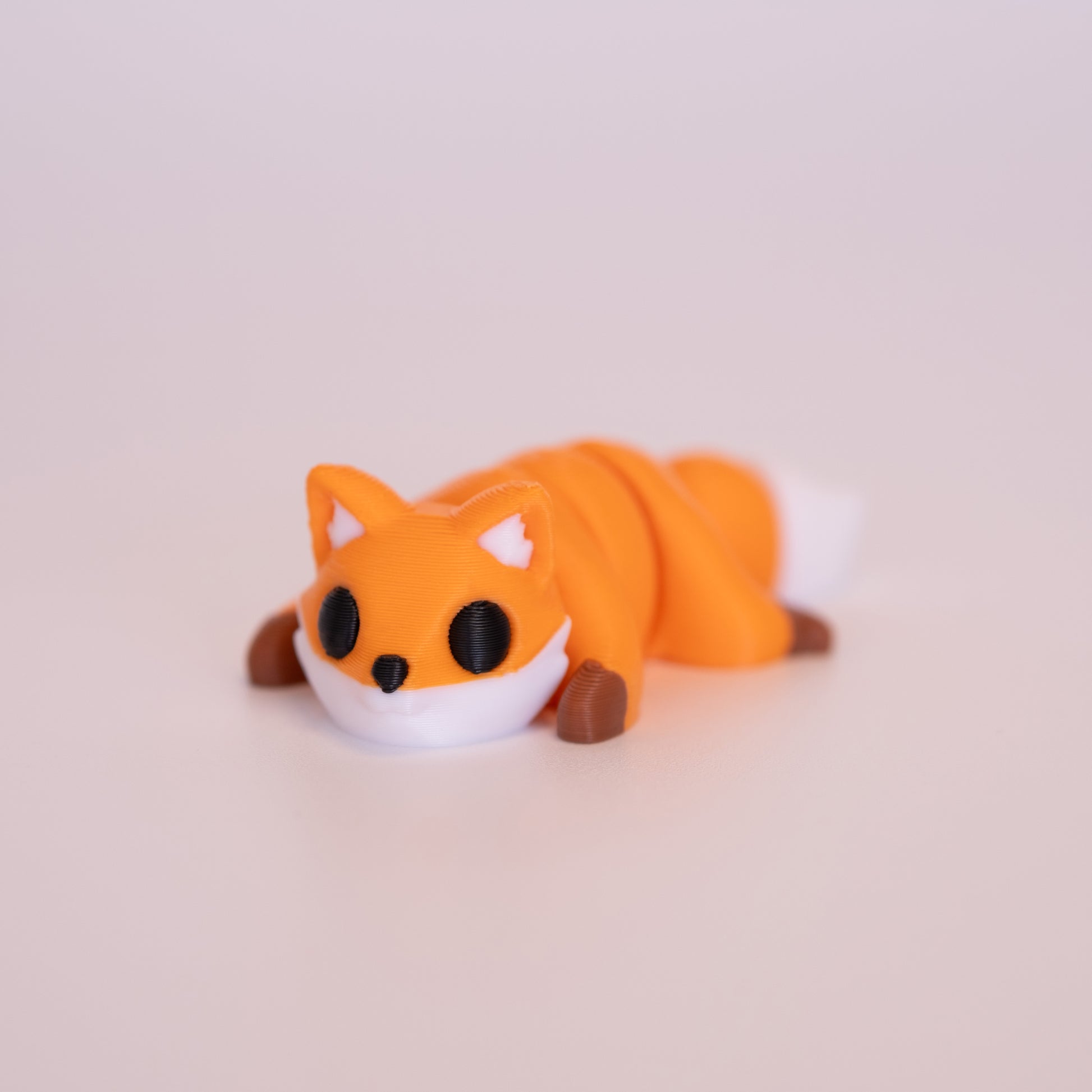 A 3D printed Red Fox figurine laying on its stomach over an off white background. The figurine is mostly orange with round black eyes, white accents, and brown paws.