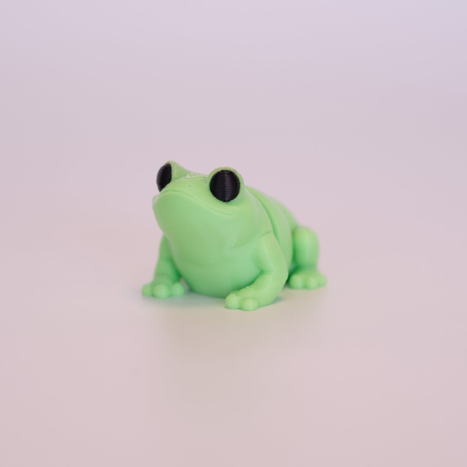 A 3D Printed Frog figurine standing on its 4 legs on an off white background. The Frog is mostly light green with round black eyes. 