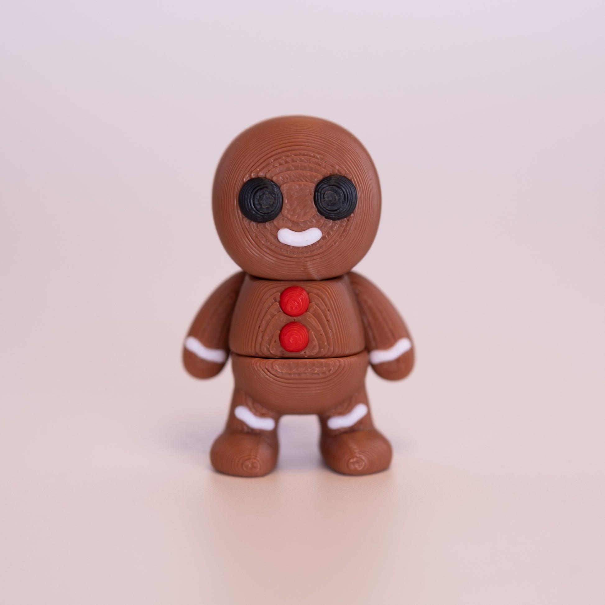 A 3D printed standing Gingerbread Man figurine on an off white background. The Gingerbread Man is a mostly warm brown, with round black eyes, red buttons, white smile, and additional white accents.