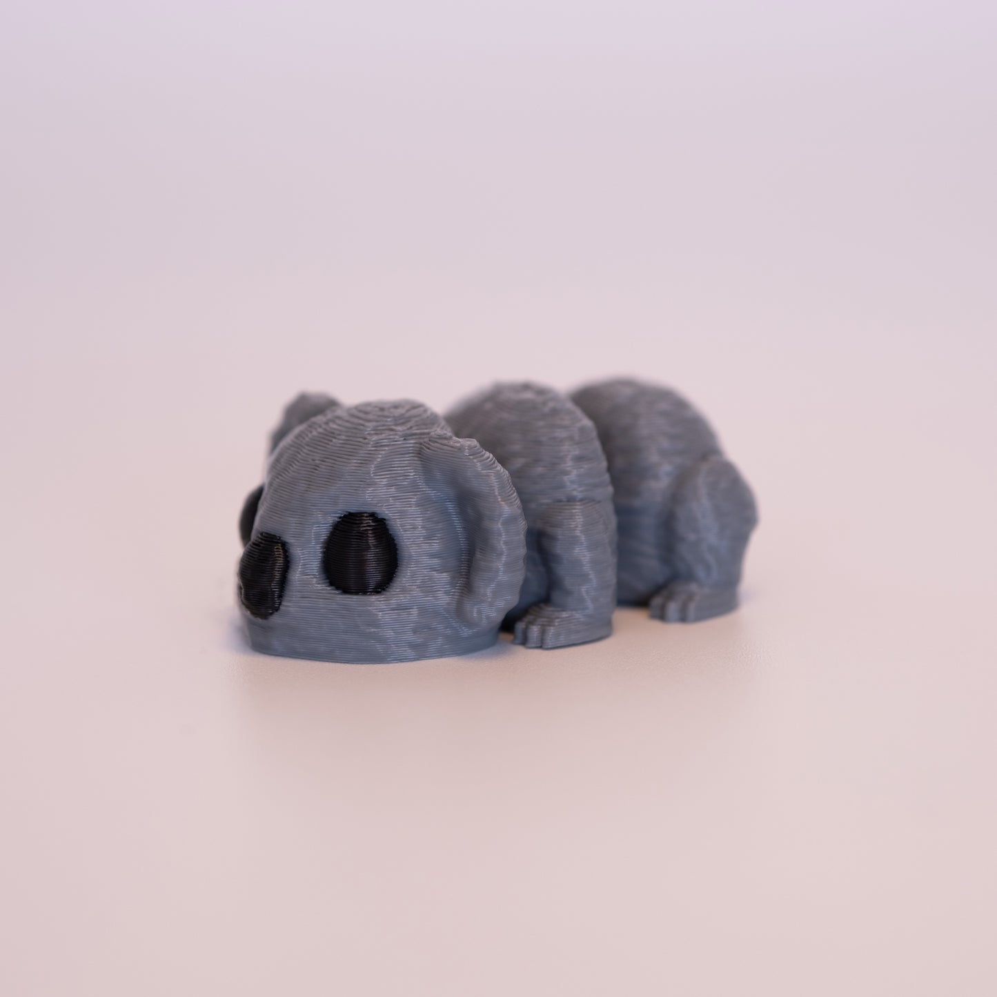 A 3D printed Koala figurine laying on its stomach over an off white background. The figurine is entirely grey with round black eyes and a black nose.