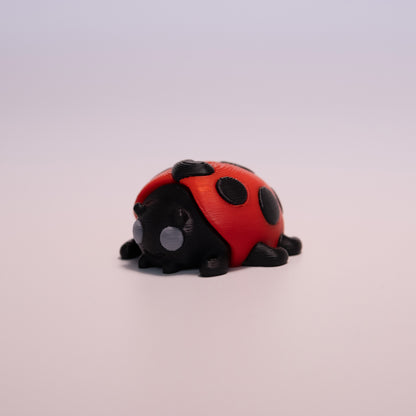 A 3D printed LadyBug figurine laying on its stomach over an off white background. The figurine is mostly black with round grey eyes, red wings, and black accent dots.