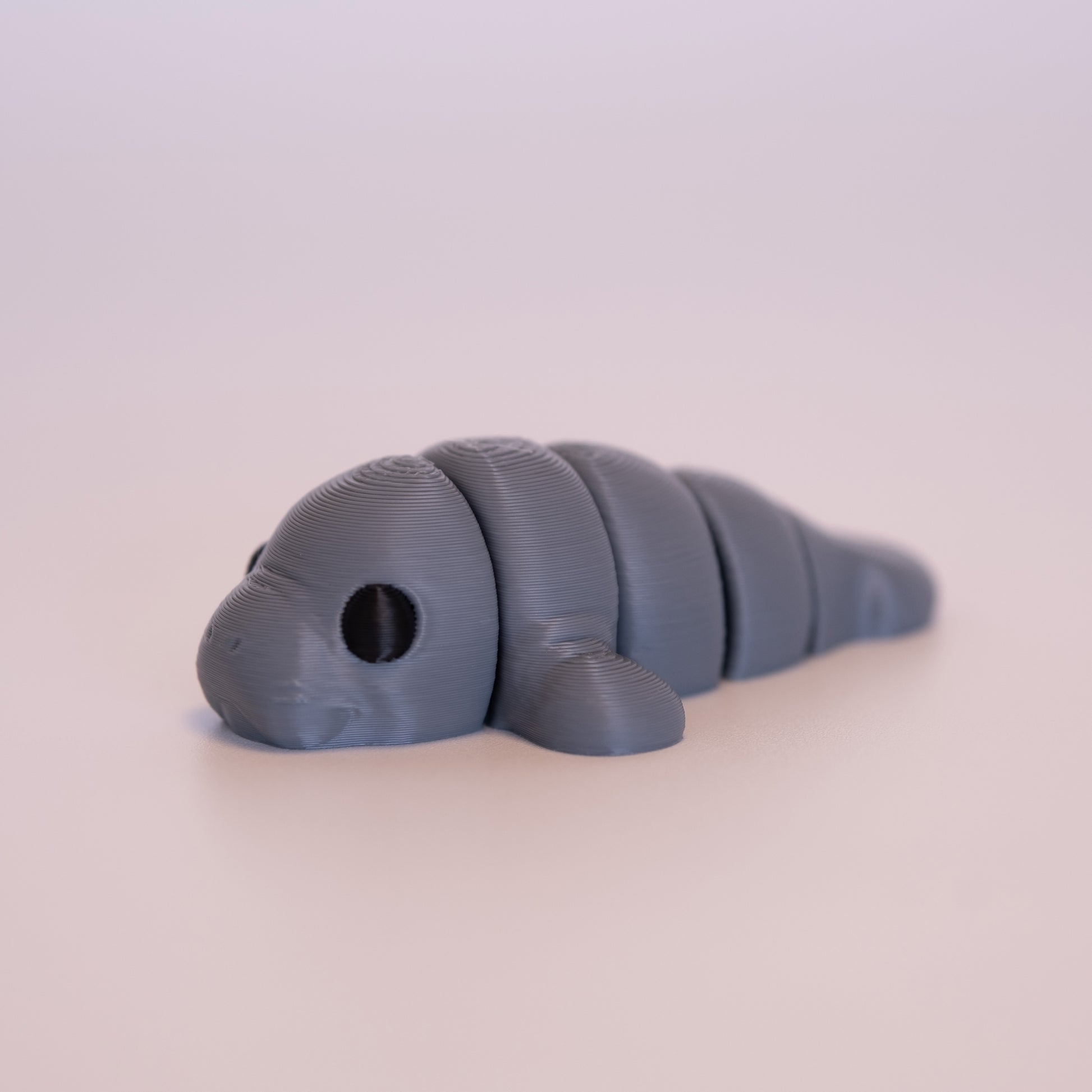 A 3D printed Manatee figurine laying on its stomach over an off white background. The figurine is entirely grey with round black eyes.