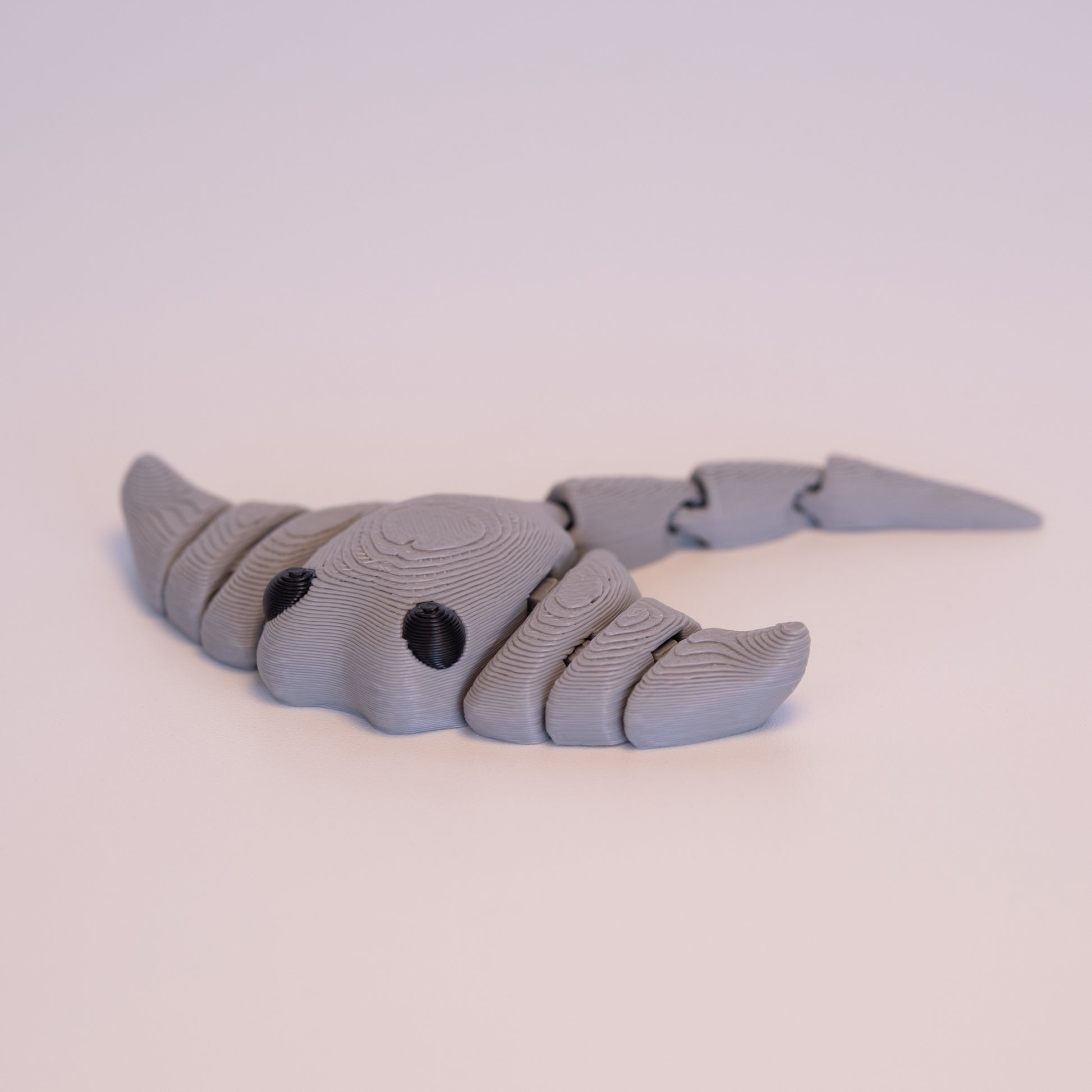 A 3D printed Manta Ray figurine laying flat on an off white background. The figurine is entirely light grey with round black eyes.