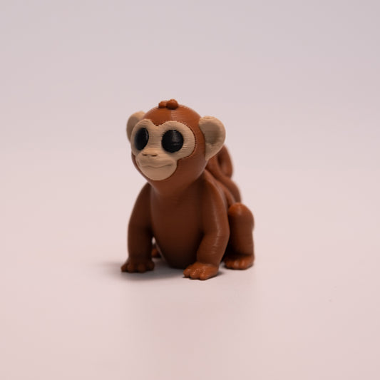 A 3D Printed Monkey figurine standing on its 4 legs on an off white background. The Monkey is mostly a warm brown with round black eyes, and tan accents on the face and ears. 