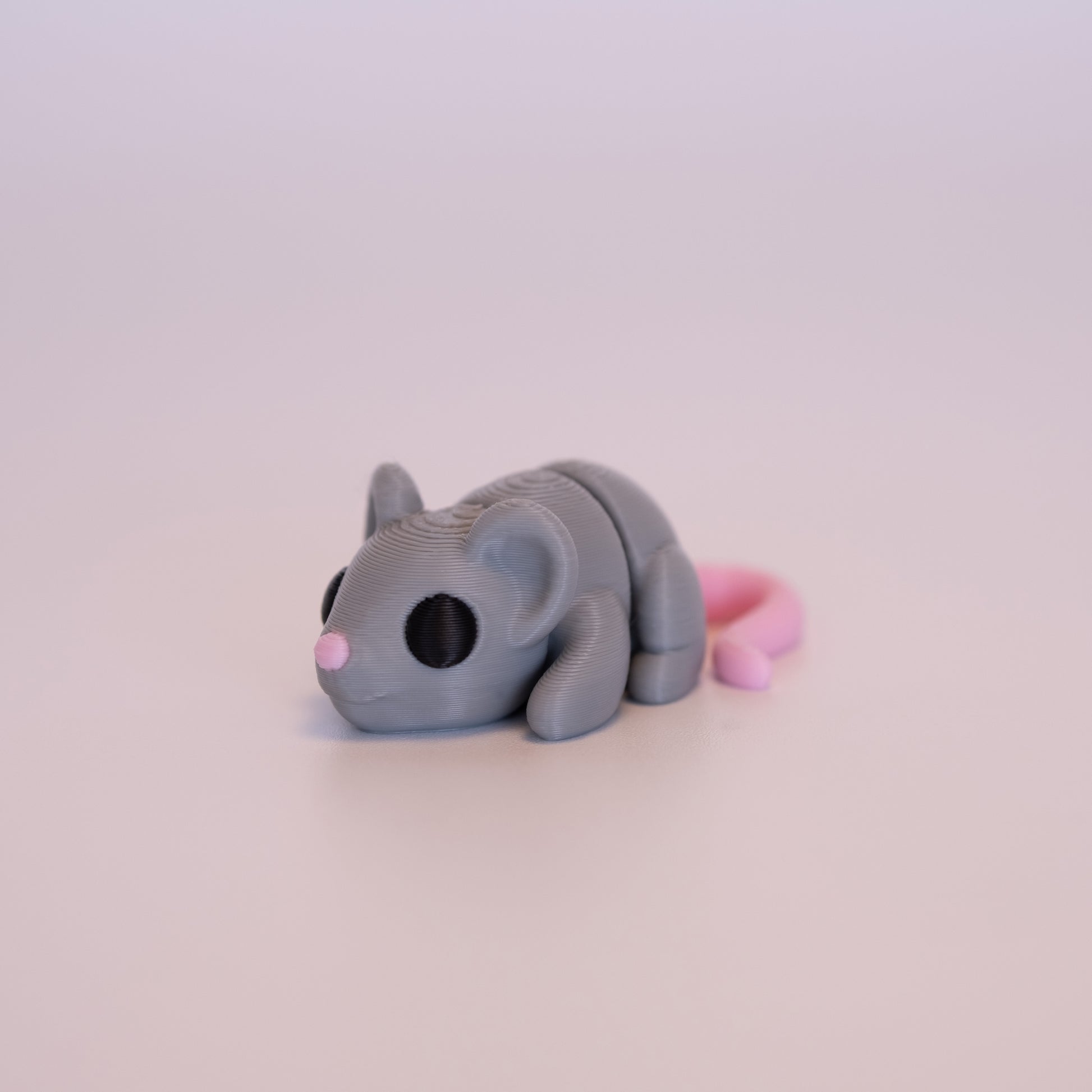A 3D printed Mouse figurine laying on its stomach over an off white background. The figurine is mostly light grey with round black eyes, pink tail, and pink nose.