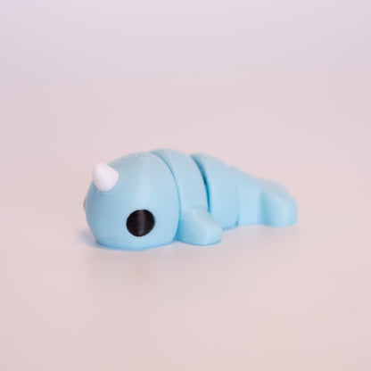 A 3D printed Narwhal figurine laying on its stomach over an off white background. The figurine is mostly light blue with round black eyes and a white horn.