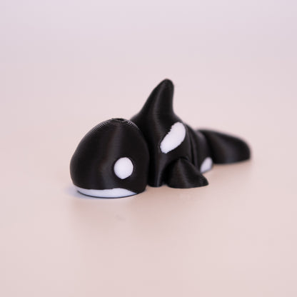 A 3D printed Orca figurine laying on its stomach over an off white background. The figurine is mostly black with round white eyes, and white accents.
