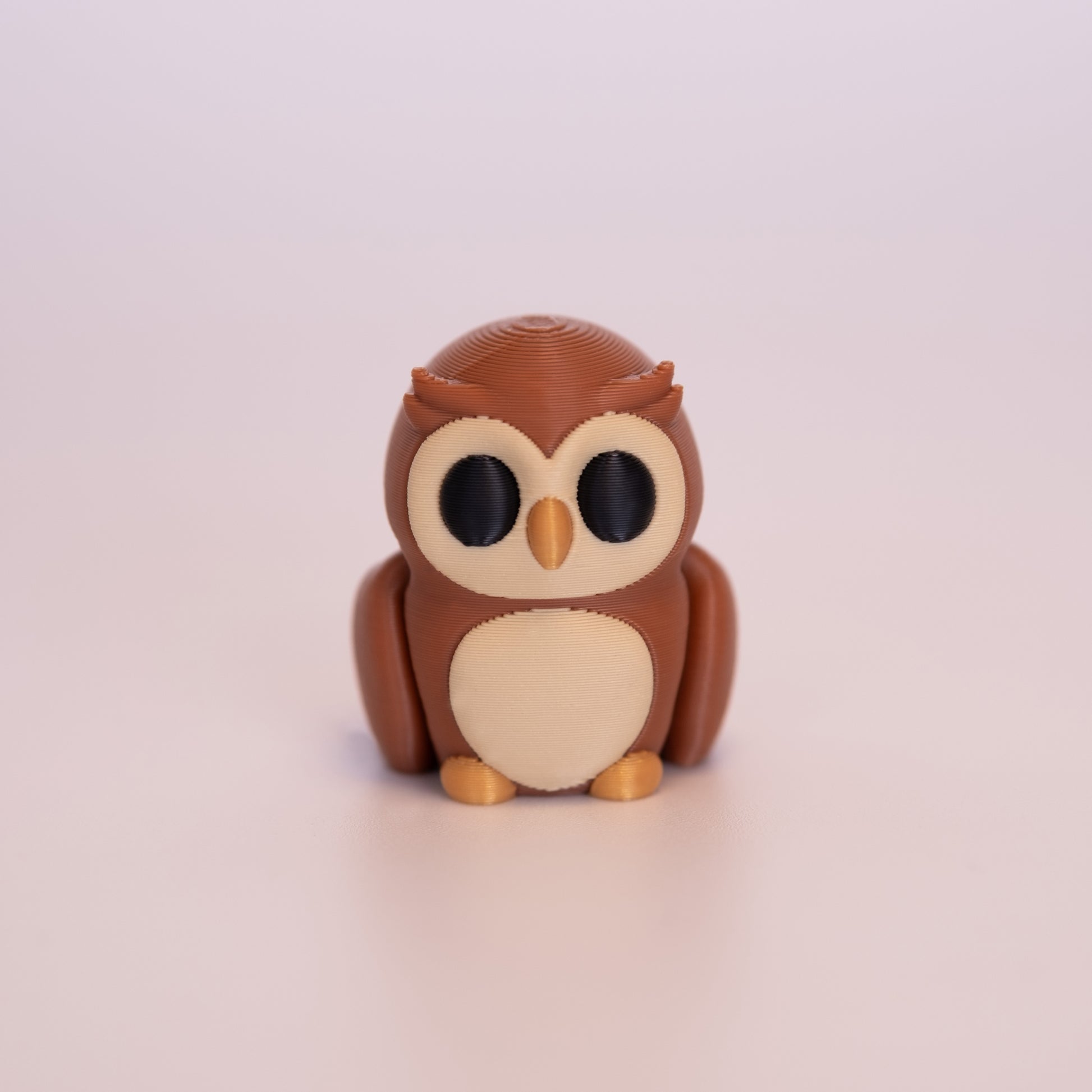A 3D printed Owl sitting upright on an off white background. The Owl is mostly a warm brown, with round black eyes, beige accents on the face and stomach, and a gold beak and feet.
