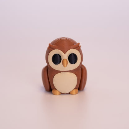 A 3D printed Owl sitting upright on an off white background. The Owl is mostly a warm brown, with round black eyes, beige accents on the face and stomach, and a gold beak and feet.